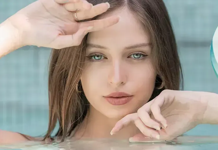Woman in water, looking at the camera, hands near her face. Brown hair, blue eyes, neutral expression.