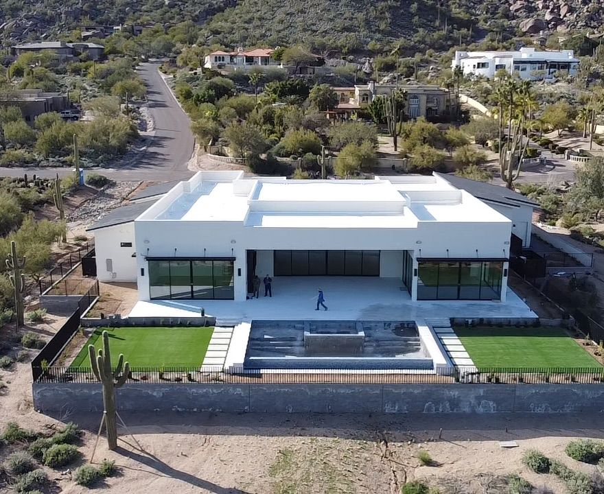 An aerial view of a modern white house with a flat roof, large glass windows, and a backyard pool in a desert landscape.
