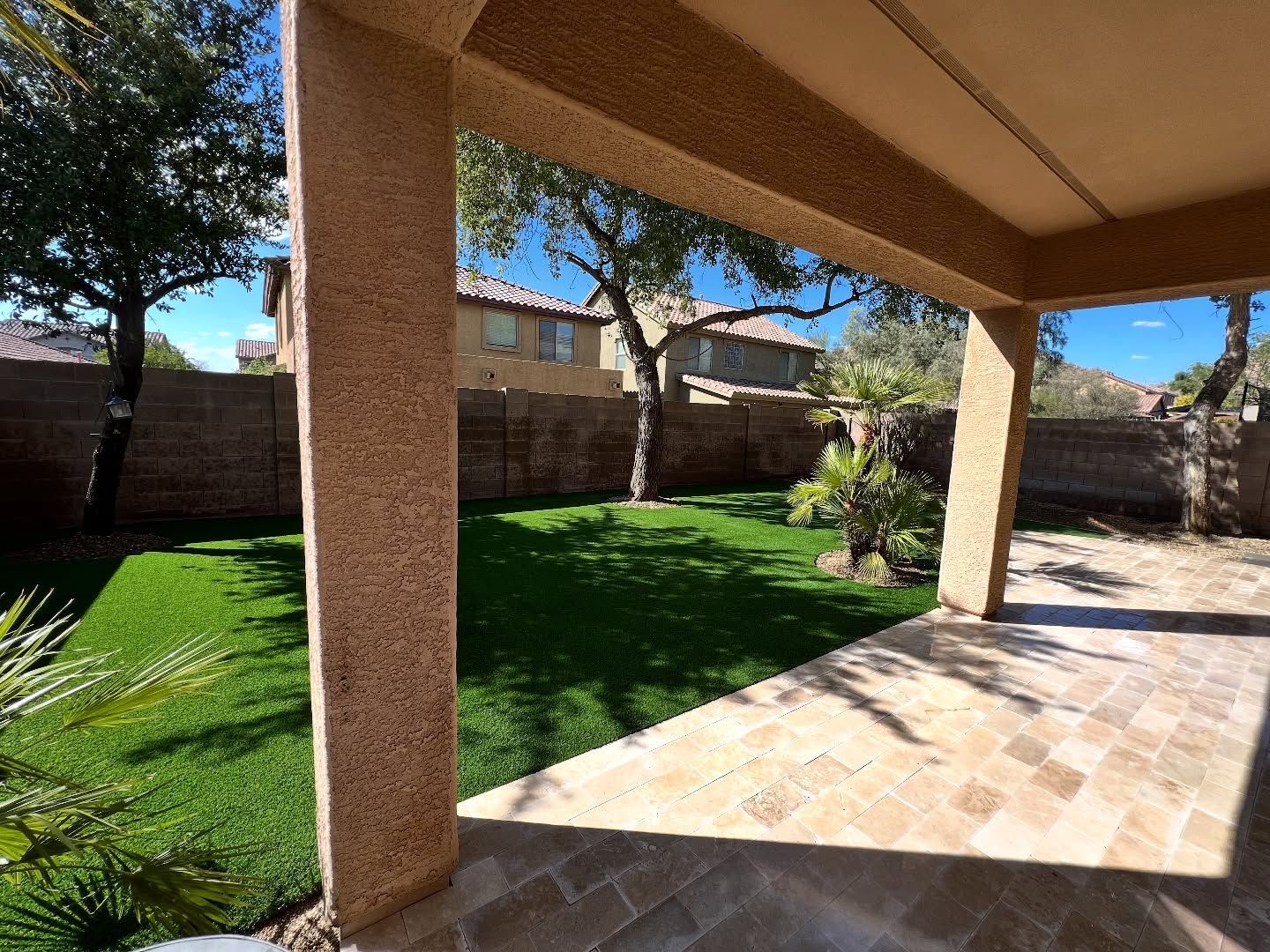 A stone-paved patio under a roof overlooks a grassy backyard with trees, a small palm, and a residential brick wall.