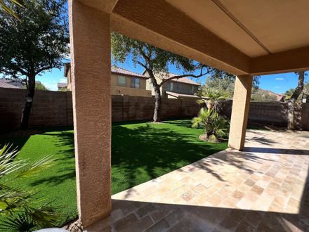 A shaded stone patio overlooks a manicured green lawn enclosed by a masonry wall and bordered by trees under a blue sky.