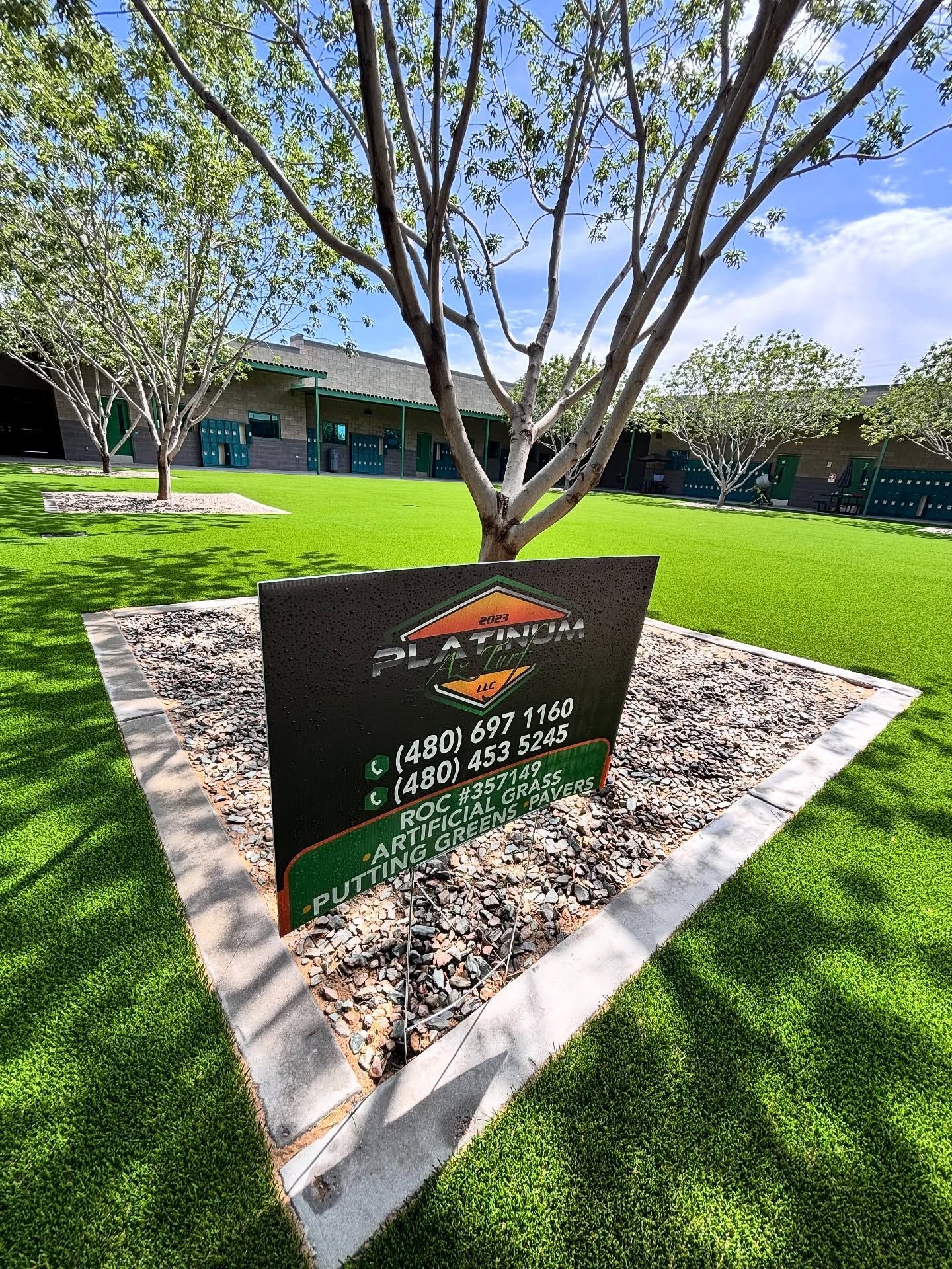 A black Platinum Turf sign sits on a gravel bed in a green lawn in front of a building under a bright blue sky.