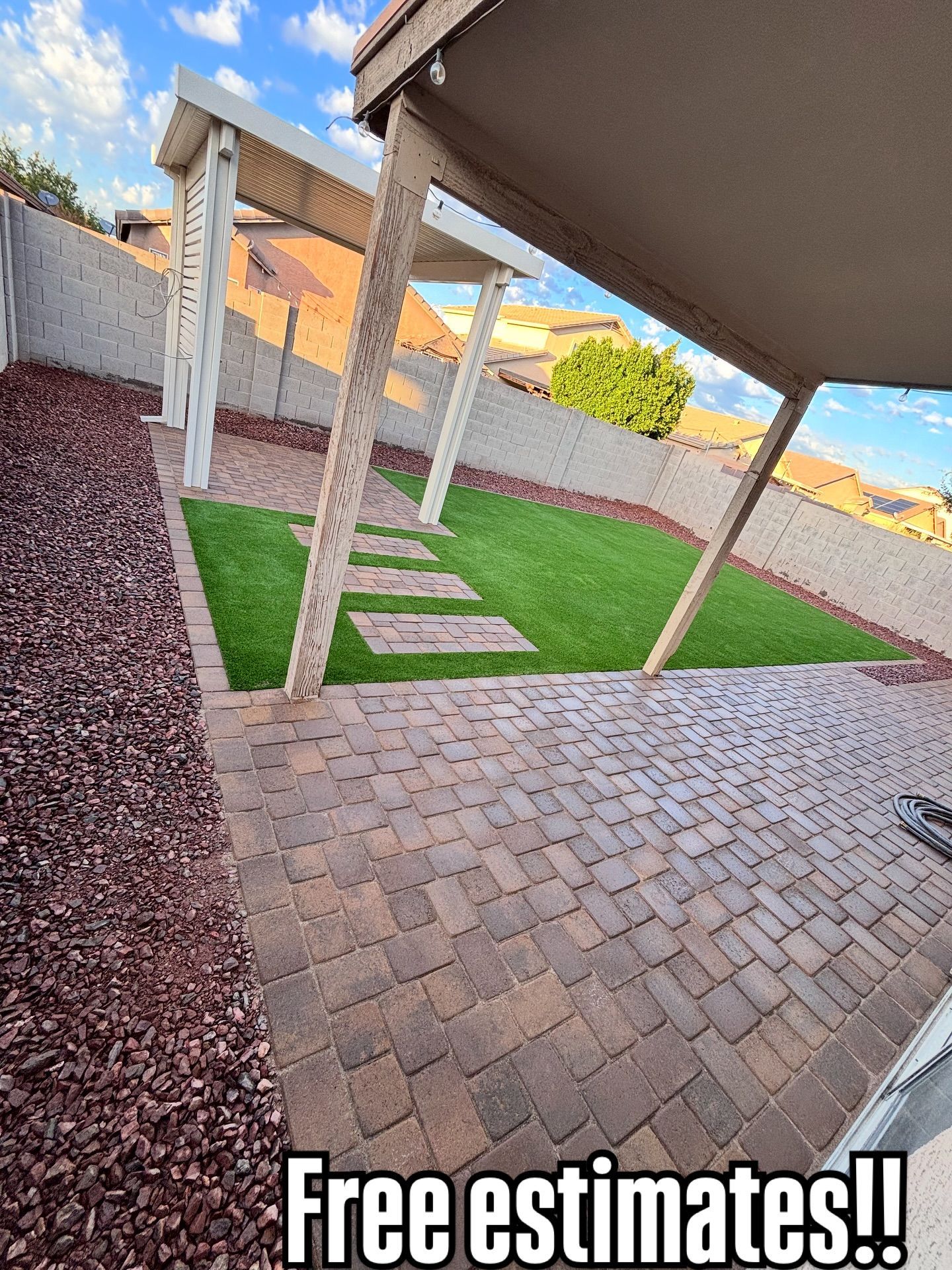 A paved patio area with artificial turf, stepping stones, and a pergola, bordered by decorative rocks and a wall.