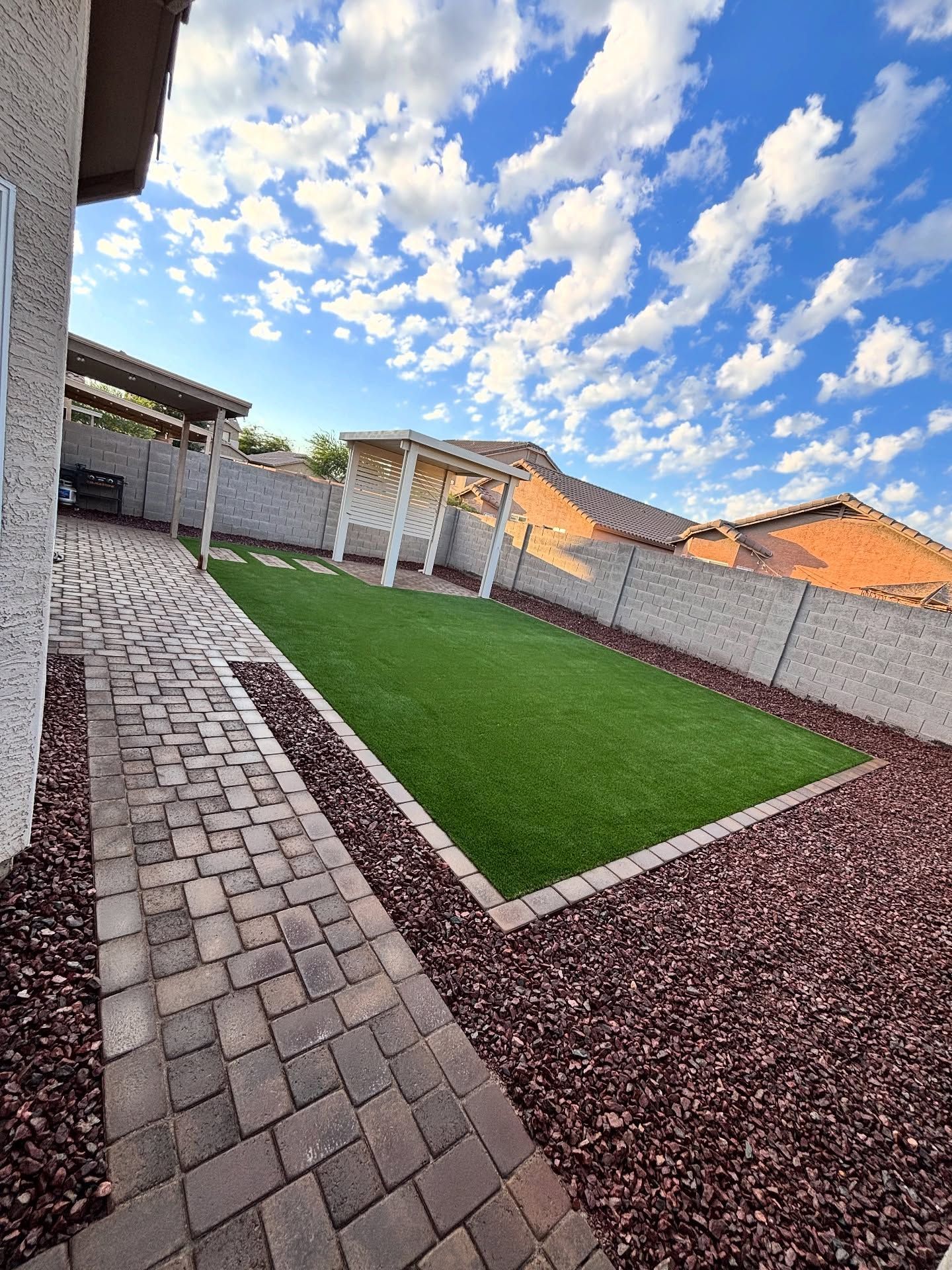 A modern backyard features a stone paver walkway, a rectangular patch of artificial green grass, and red gravel accents.