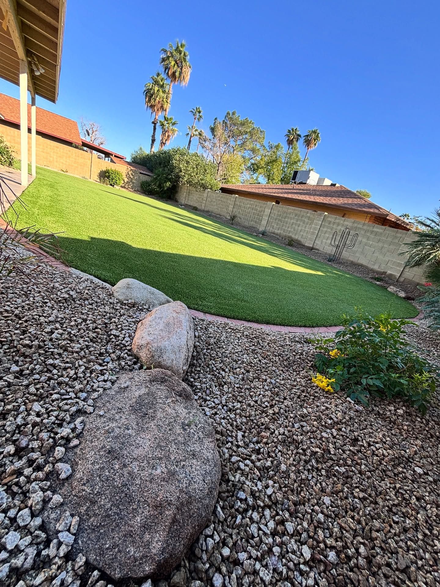 A sunny backyard featuring a green grass lawn, desert landscaping with gravel and rocks, and palm trees against a blue sky.