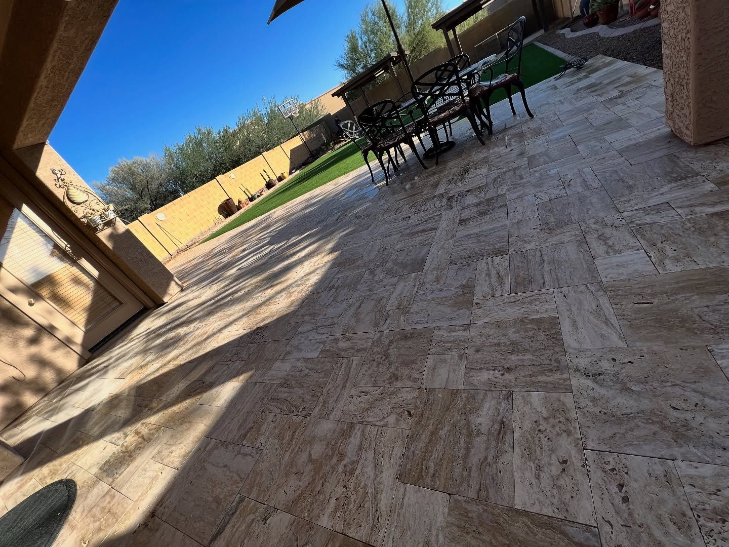 A tan stone patio featuring a rectangular tiling pattern leads toward a small outdoor table and chairs in a backyard.