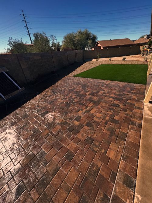 A backyard featuring a large, multi-toned brick paver patio and a rectangular patch of artificial green grass.