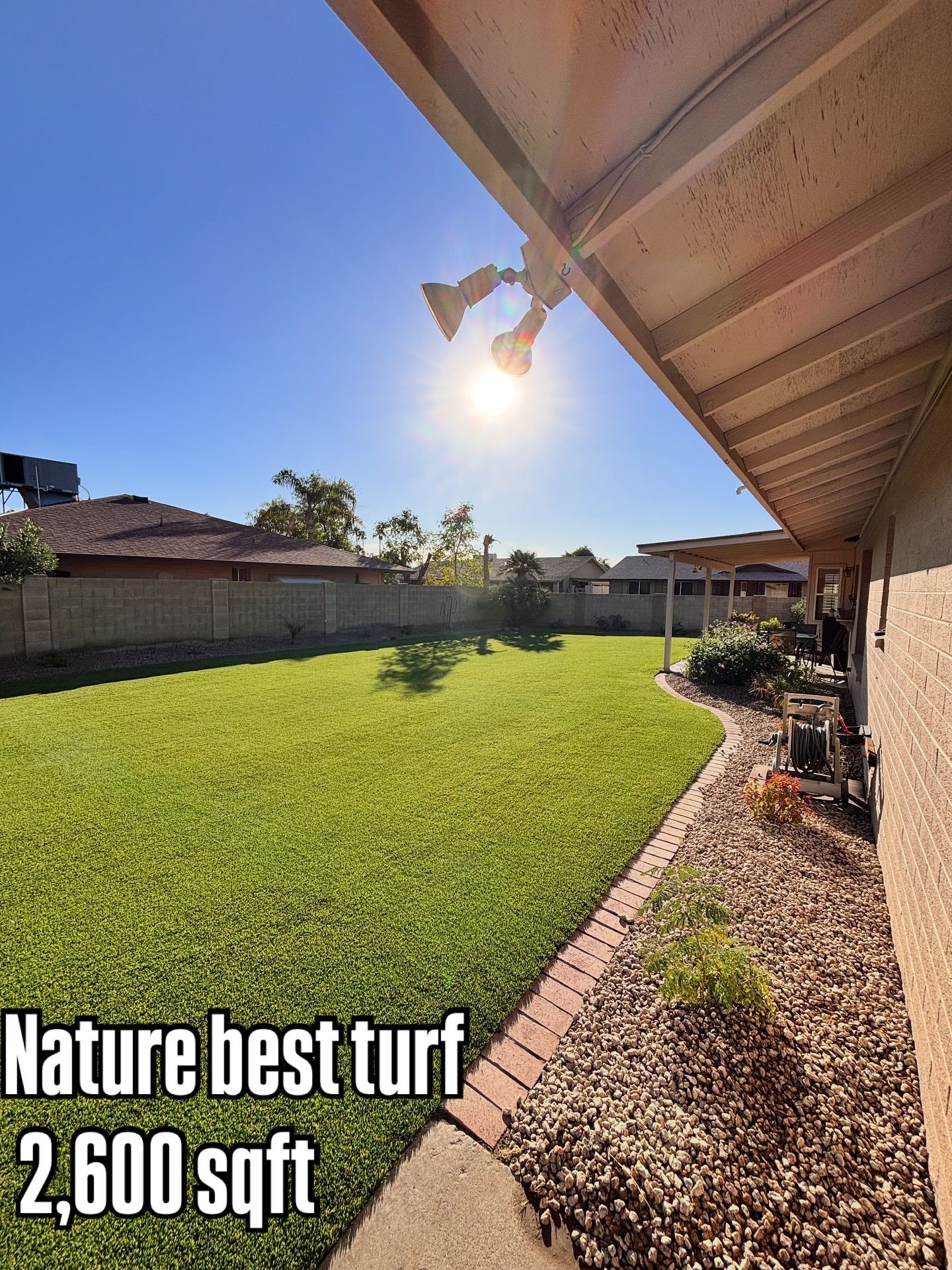 A sunlit residential backyard featuring a green turf lawn and a stone-lined garden path next to a tan brick house wall.