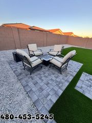 Four chairs and a square fire pit on a gray paver patio in a backyard with artificial turf and a gravel border.