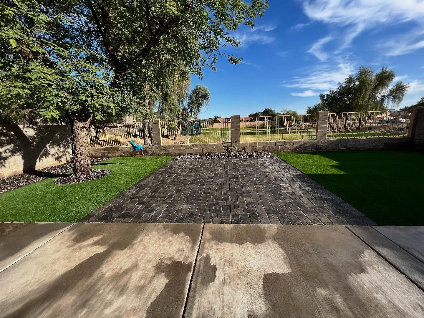 A backyard view featuring a dark stone patio bordered by artificial turf, trees, and a low wall under a sunny blue sky.