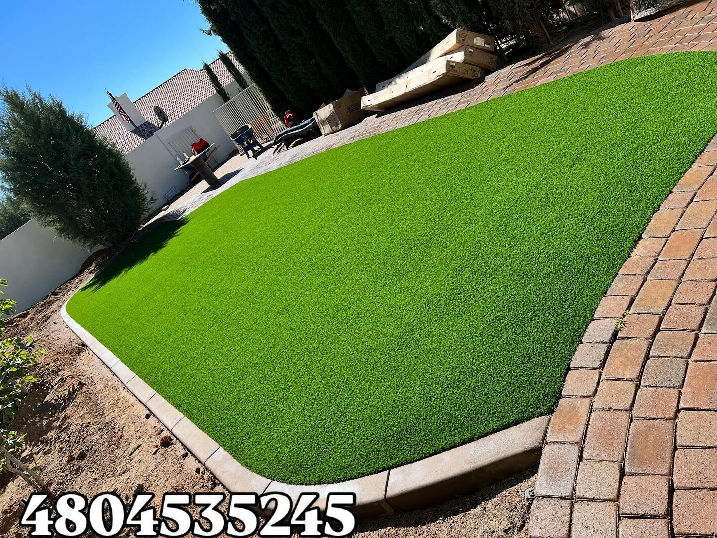 A manicured green artificial grass lawn bordered by tan stone pavers and a brick walkway in a residential backyard.