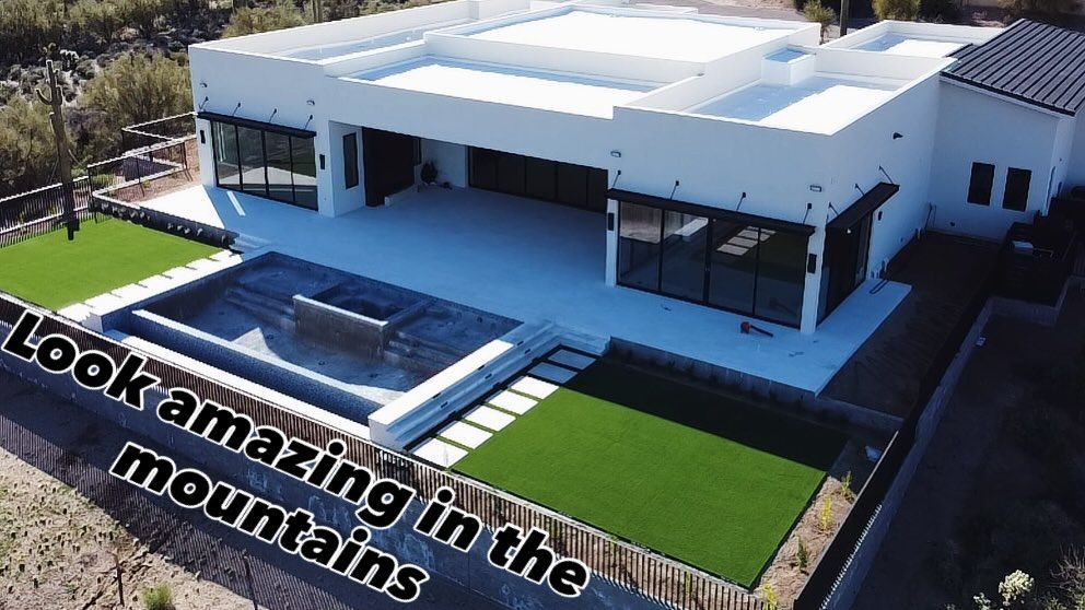 An aerial view of a modern white house with a sunken patio, swimming pool, and artificial grass lawn in a desert setting.