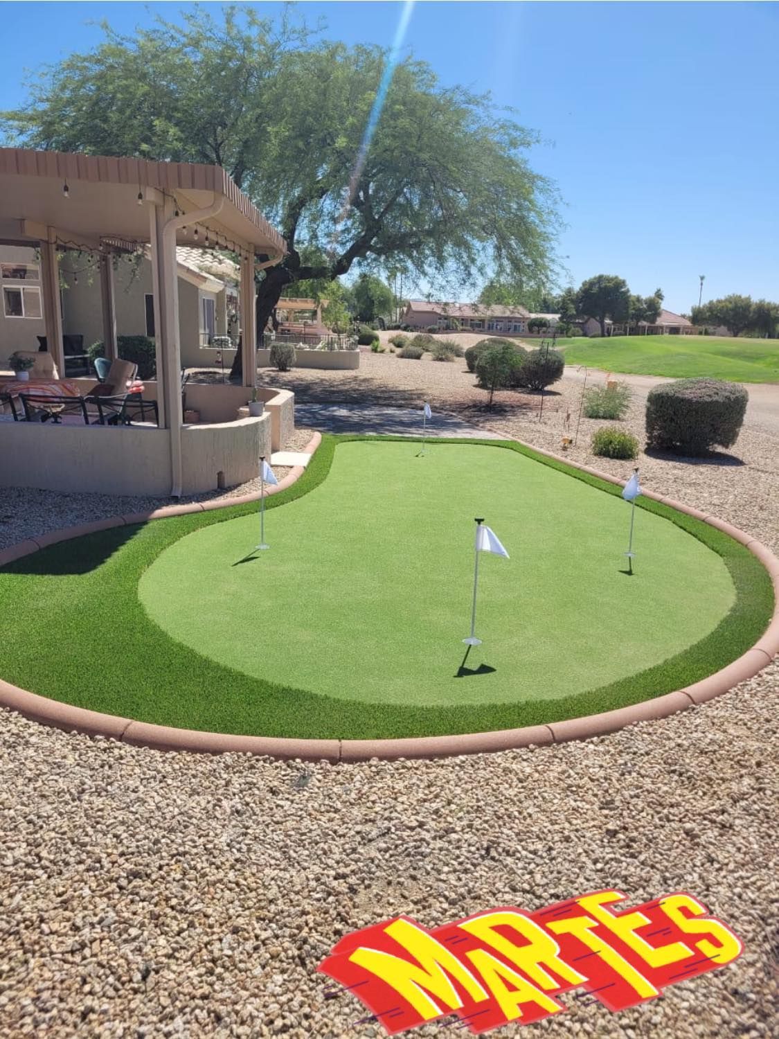 A backyard putting green with multiple holes and white flags, beside a covered patio in a sunny, desert-landscaped yard.