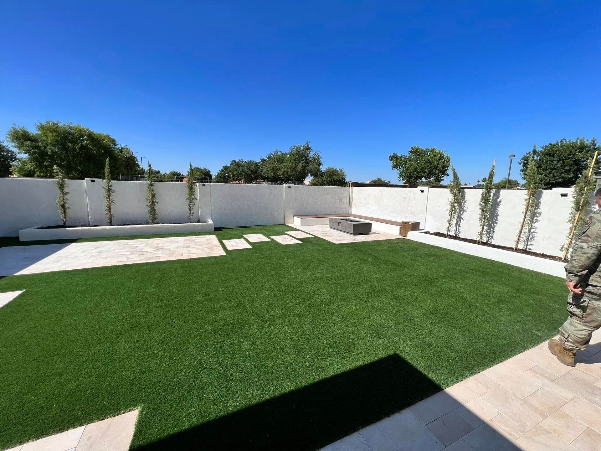 A landscaped backyard featuring artificial grass, light-colored pavers, a white wall with small trees, and a fire pit area.