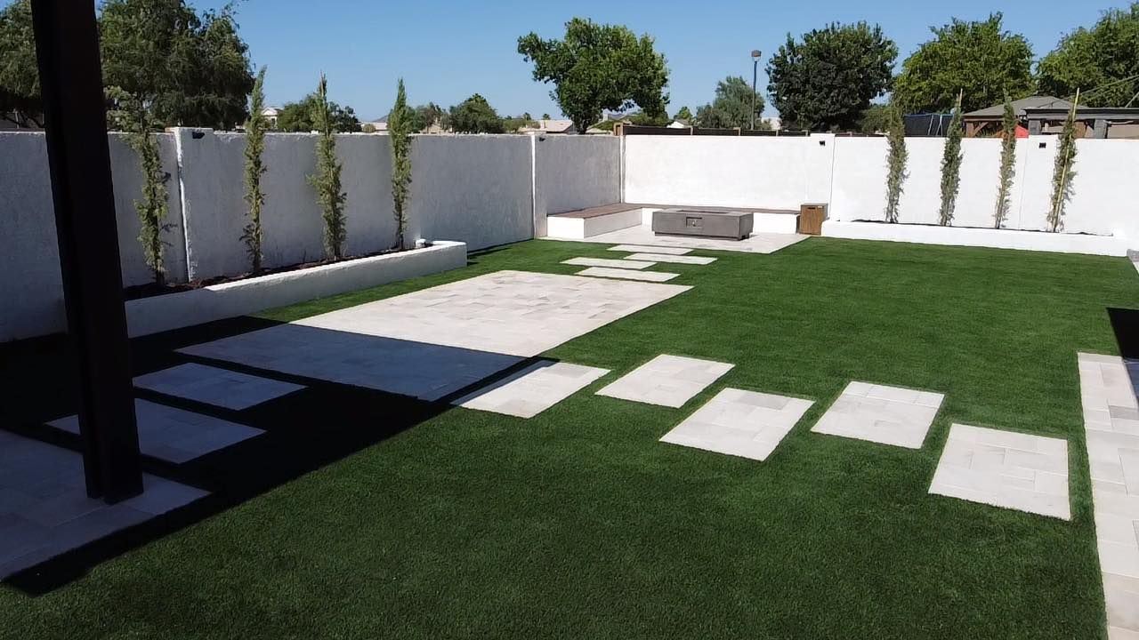 A modern backyard featuring a green lawn, stone stepping stones, a patio area, and white walls with slender trees.