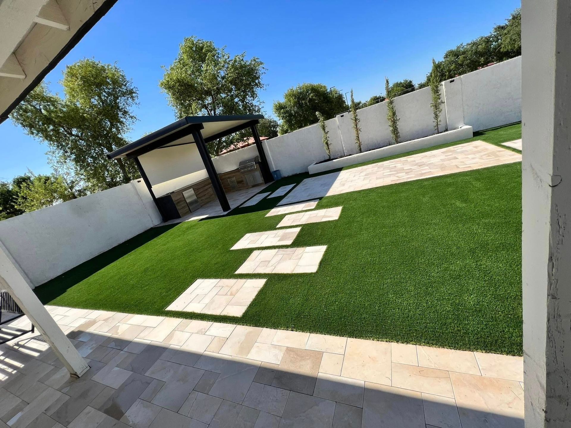 A modern backyard with artificial turf, stone pavers, a white perimeter wall, and a shaded outdoor kitchen area.