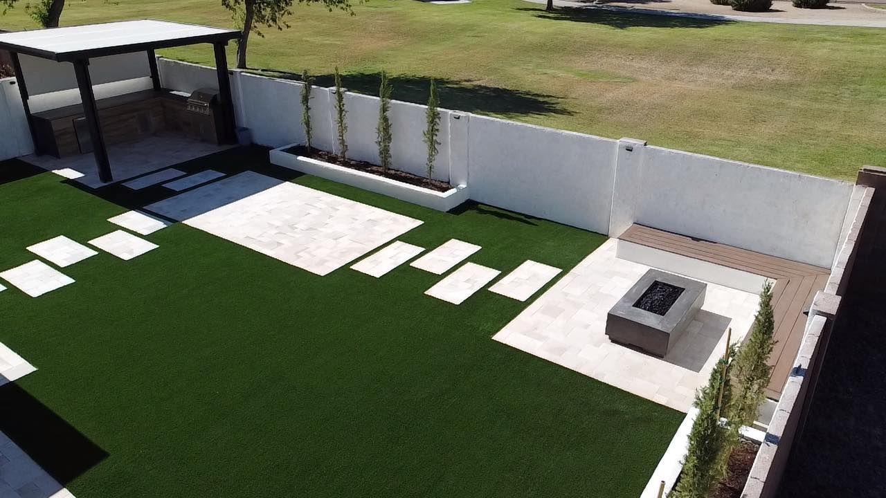 A modern backyard with synthetic turf, white paver pathways, a fire pit seating area, and a shaded pergola.