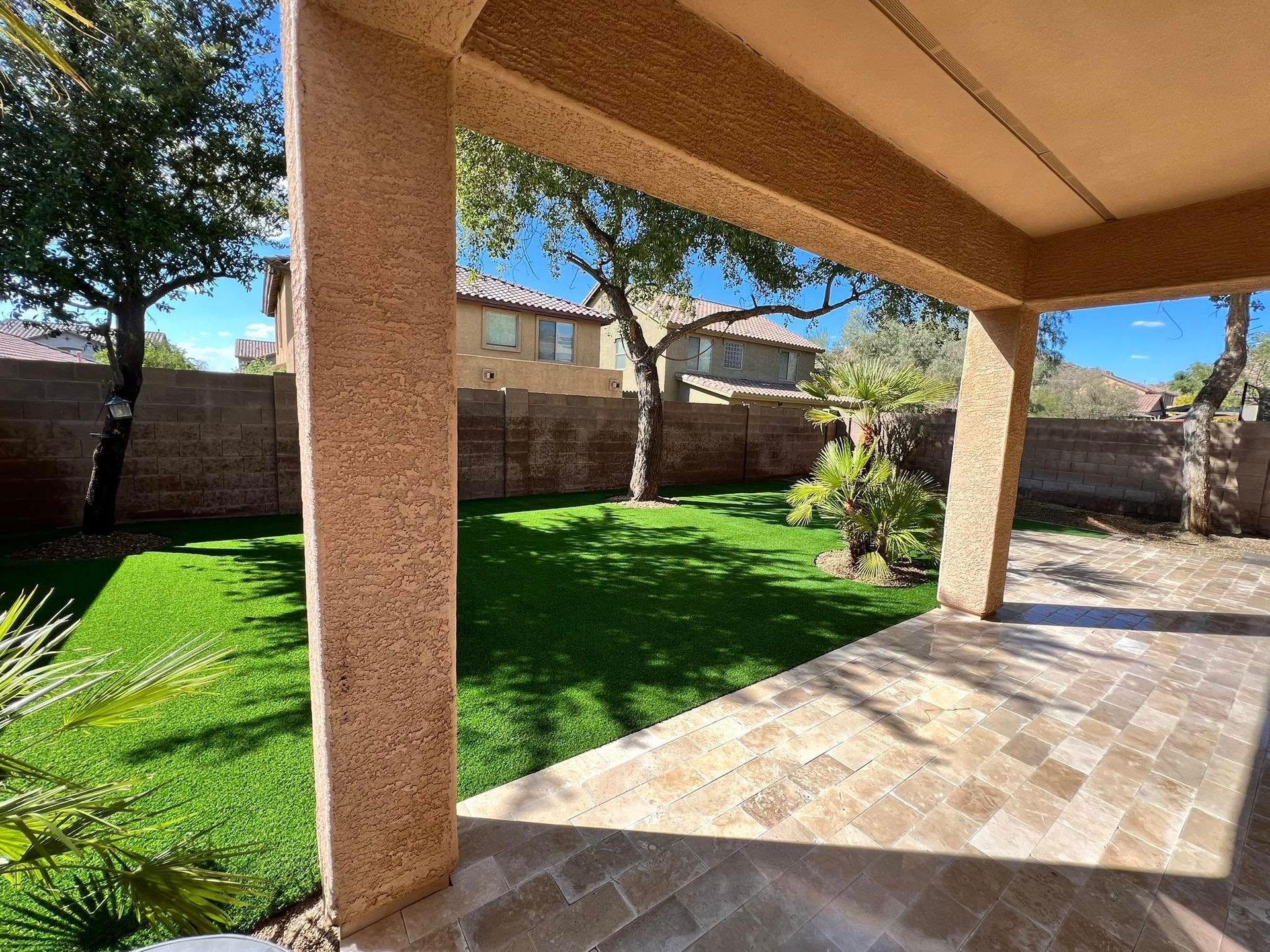 A view from a covered patio looking out onto a small grassy backyard enclosed by a brown stone wall and trees.