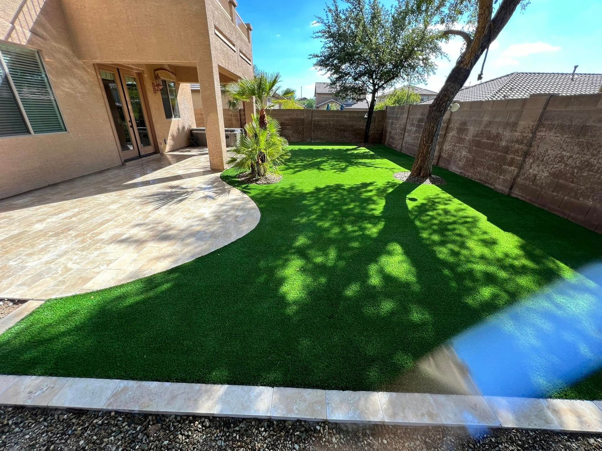 A backyard with a paved patio, green artificial turf, a small palm tree, and a block wall fence under a blue sky.