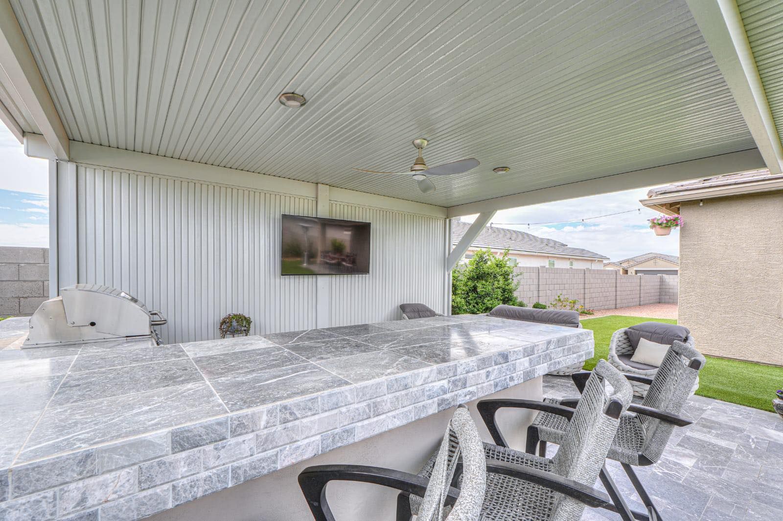 A shaded outdoor kitchen patio with a stone bar, seating, and a mounted flat-screen TV against a white metal wall.