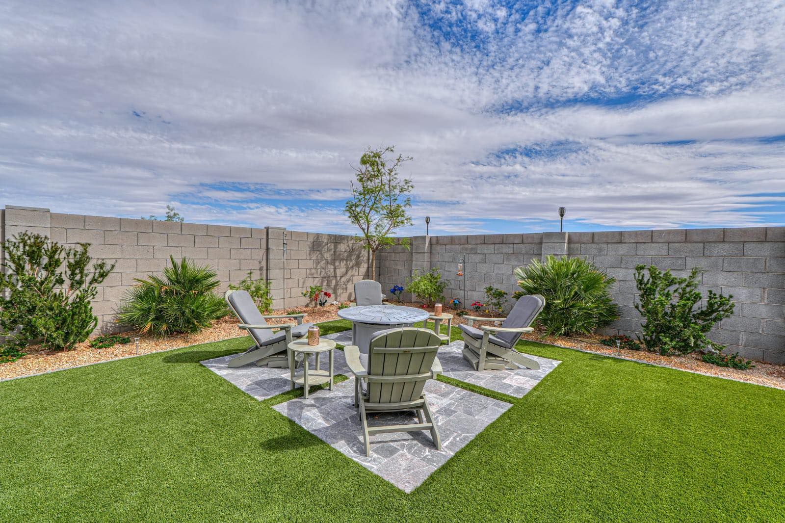 A backyard with a stone patio, a small table, and four Adirondack chairs set against a block wall under a blue sky.