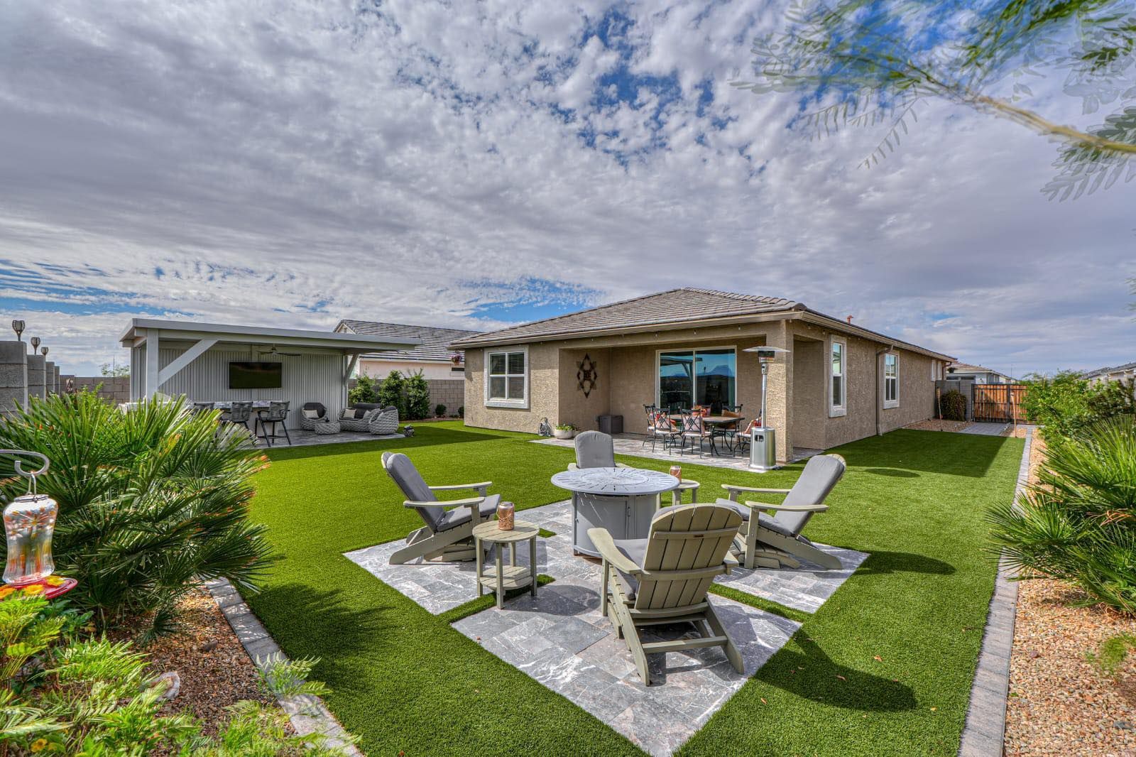 A backyard with a stone fire pit area, Adirondack chairs, a covered patio, and manicured lawn under a cloudy blue sky.