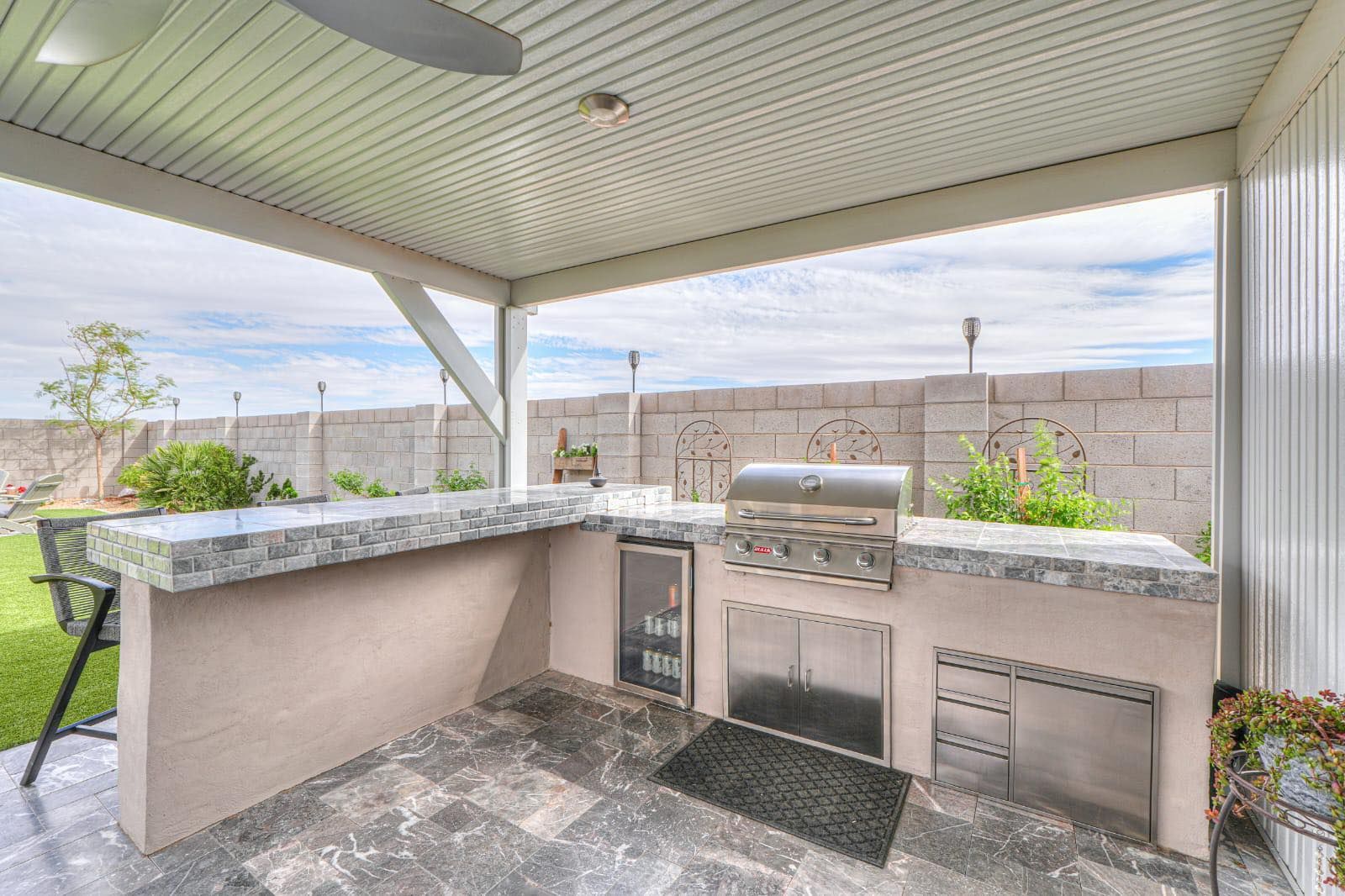 An outdoor kitchen under a covered patio with a granite-topped bar, stainless steel grill, beverage fridge, and grill.