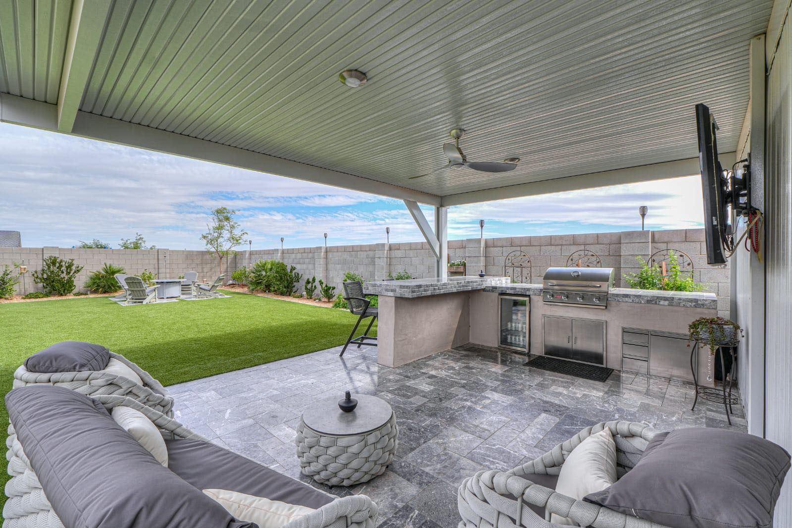 A covered outdoor patio featuring a kitchen area with a grill, gray seating, and a view of a grassy backyard.