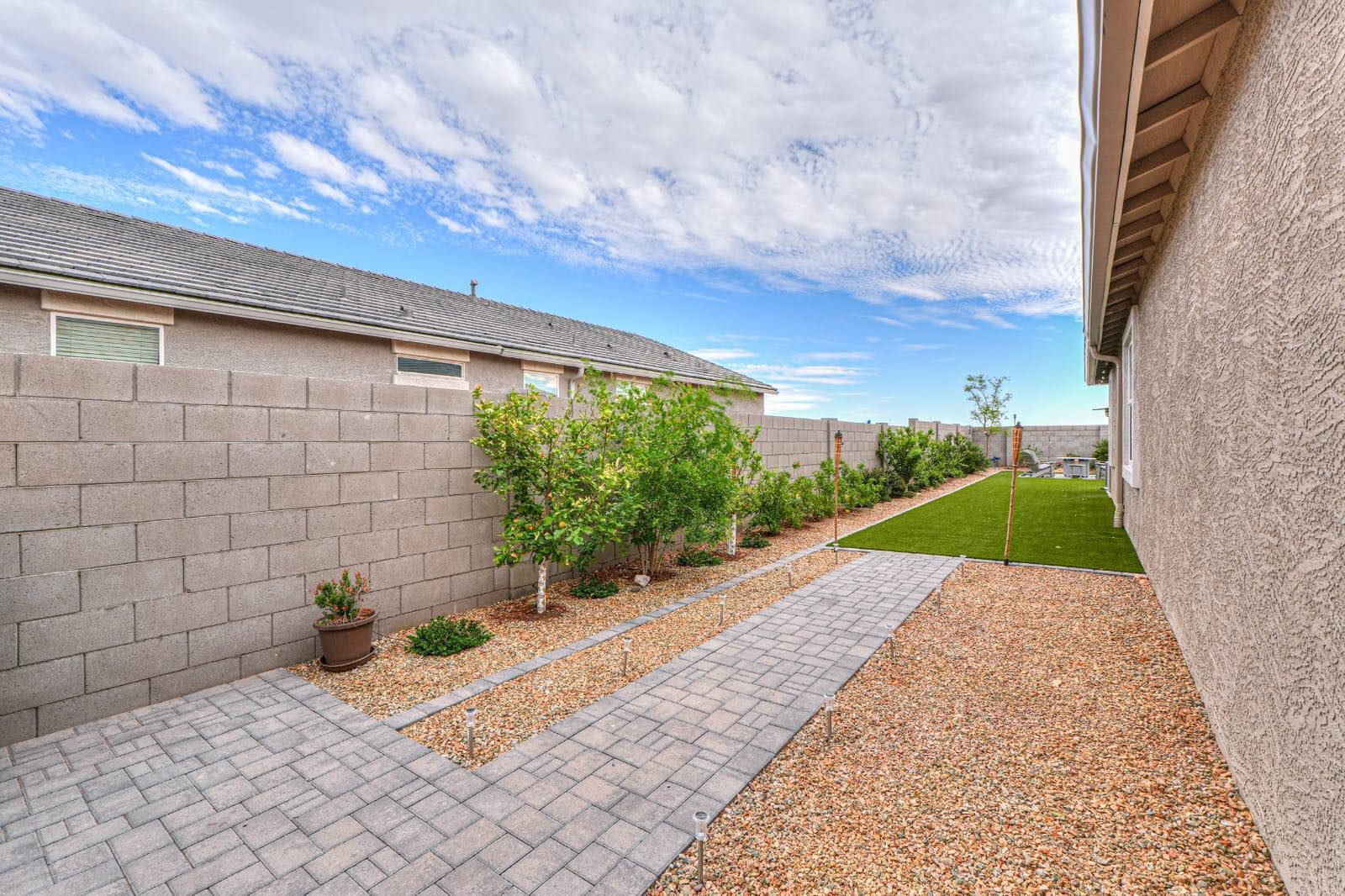 A side yard with a paver walkway, light-colored gravel, a small patch of grass, and a concrete block wall.