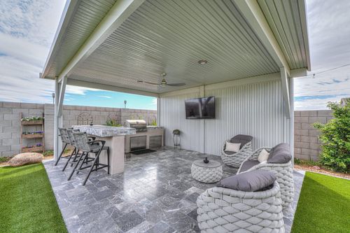 An outdoor covered patio features a kitchen island with bar stools, wicker chairs, a small table, and a mounted TV.