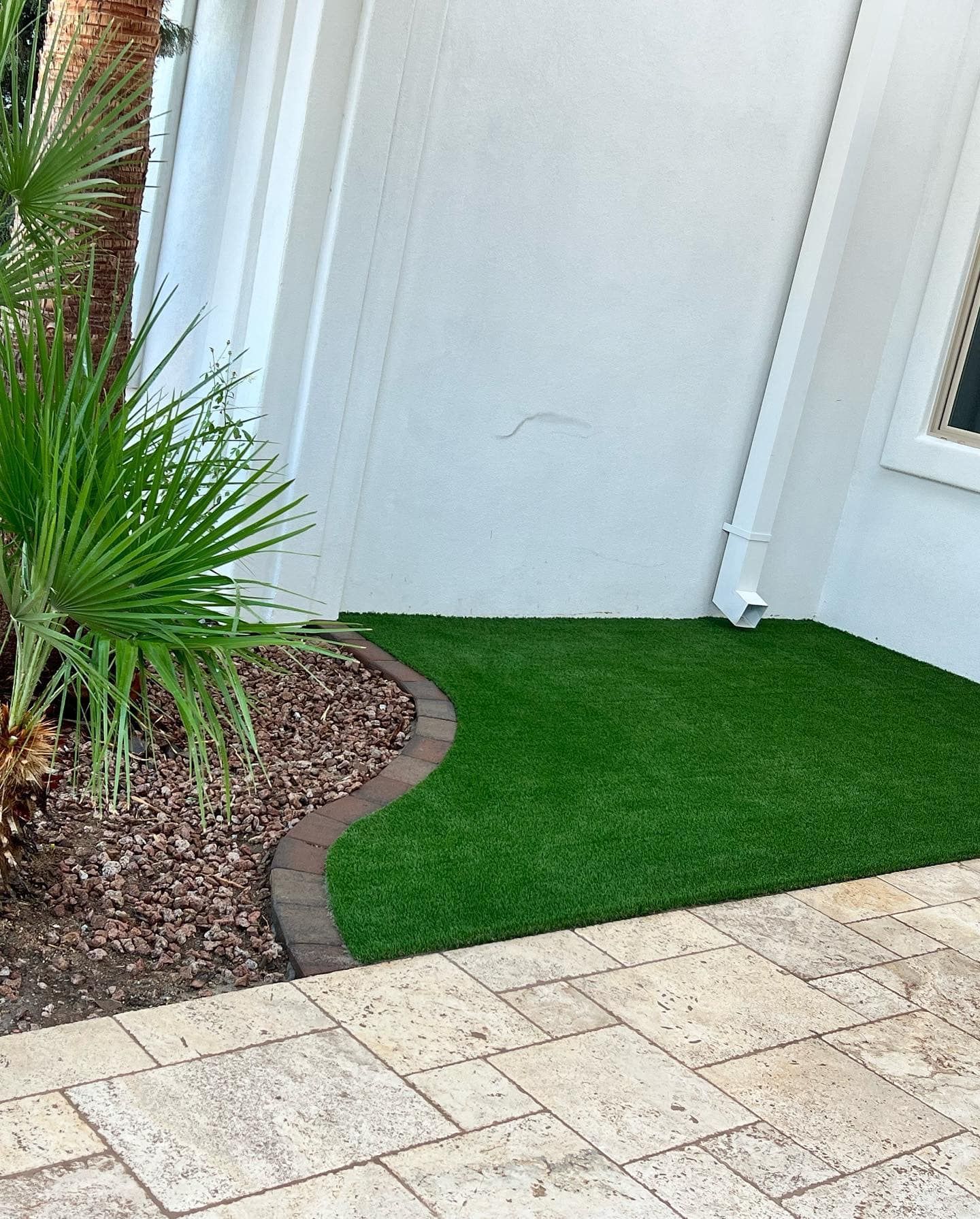 An area with artificial turf, bordered by curved pavers next to a light-colored building wall and a stone patio.