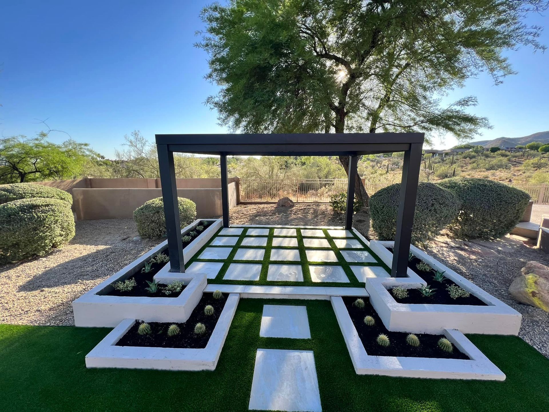 A modern outdoor pergola sits on a grid of white pavers and artificial turf, surrounded by white planters and desert views.