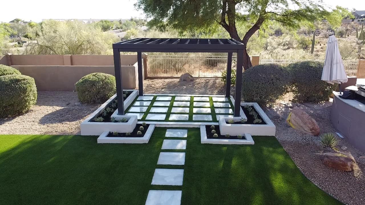 A dark pergola stands over a grid of stone pavers and synthetic grass in a backyard, linked by a stone path to the lawn.