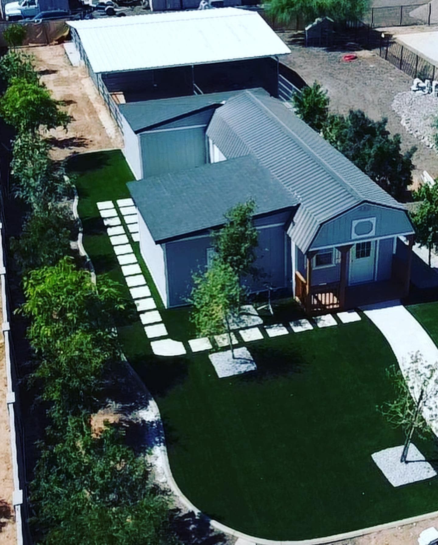 An aerial view of a gray house with a green roof, manicured lawn, stone pathway, and an adjacent carport or structure.