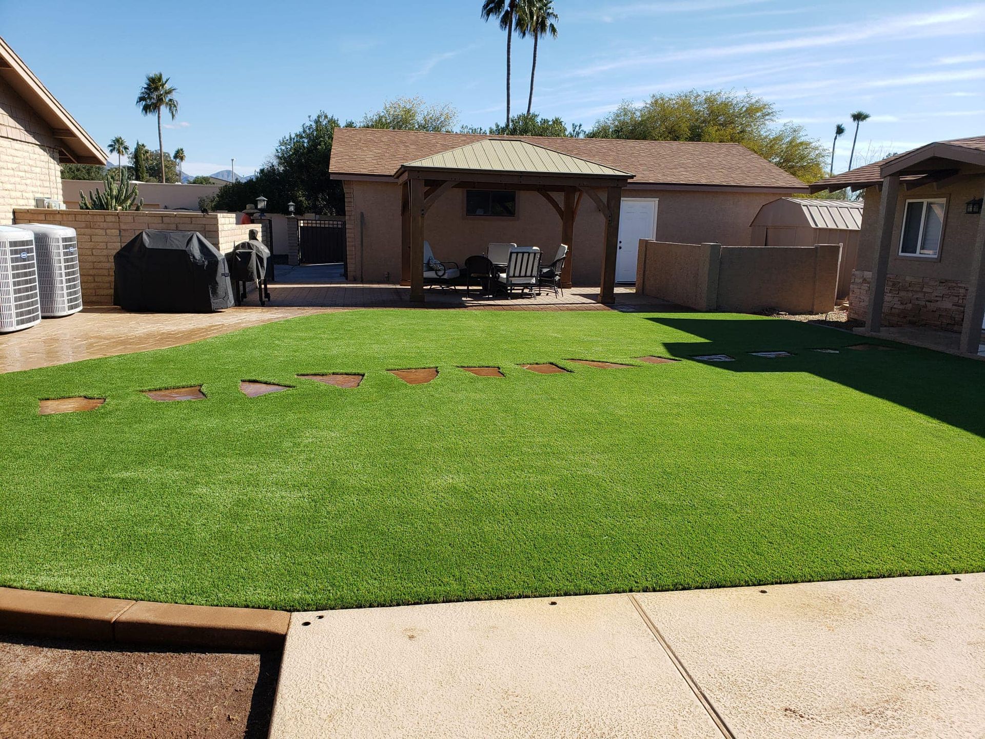 A sunny backyard with a lush green artificial lawn, a stone paver path, a covered patio area, and a beige stucco house.