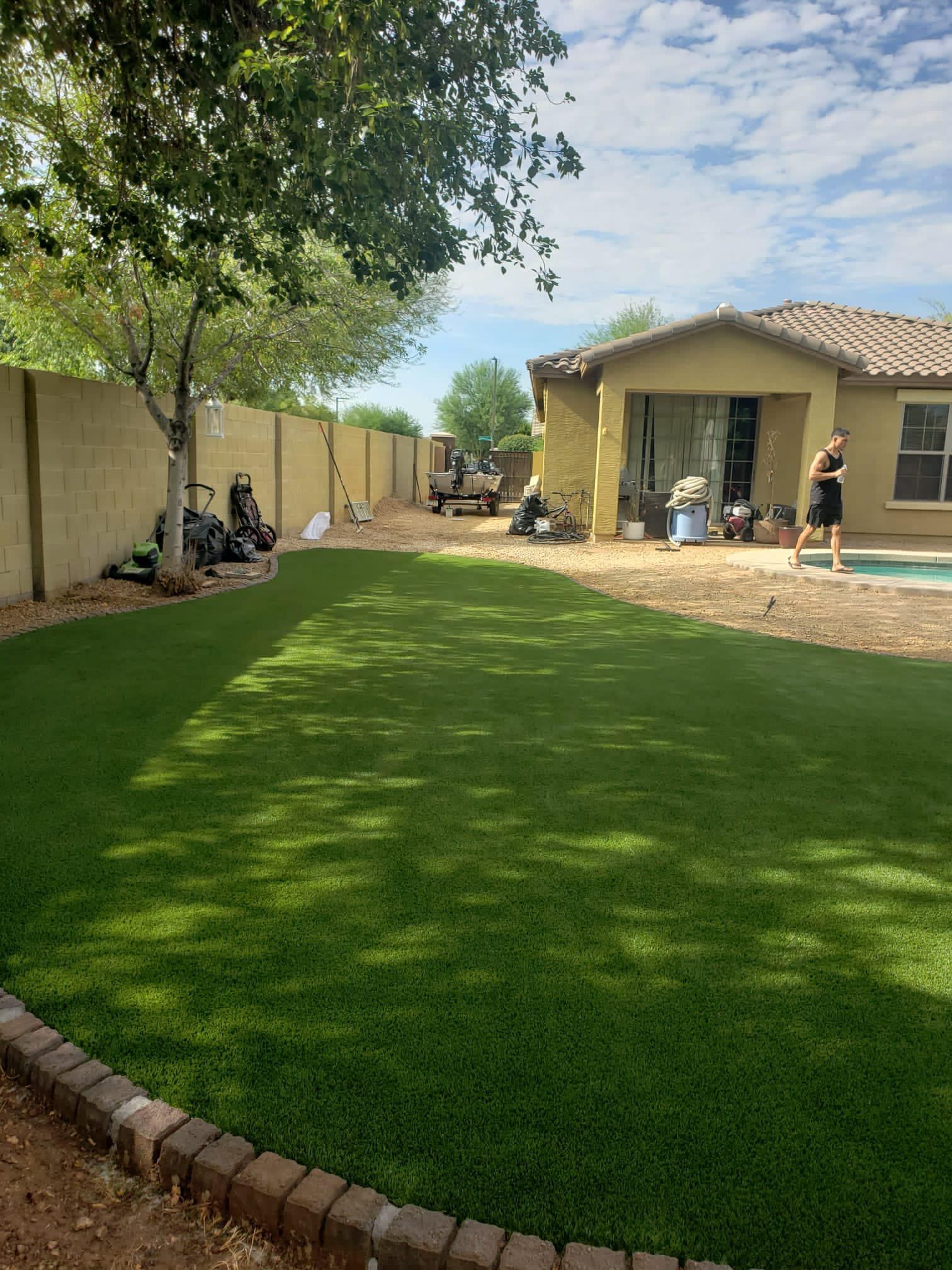 A bright green artificial turf lawn in a suburban backyard, bordered by bricks with a house and patio area in the background.