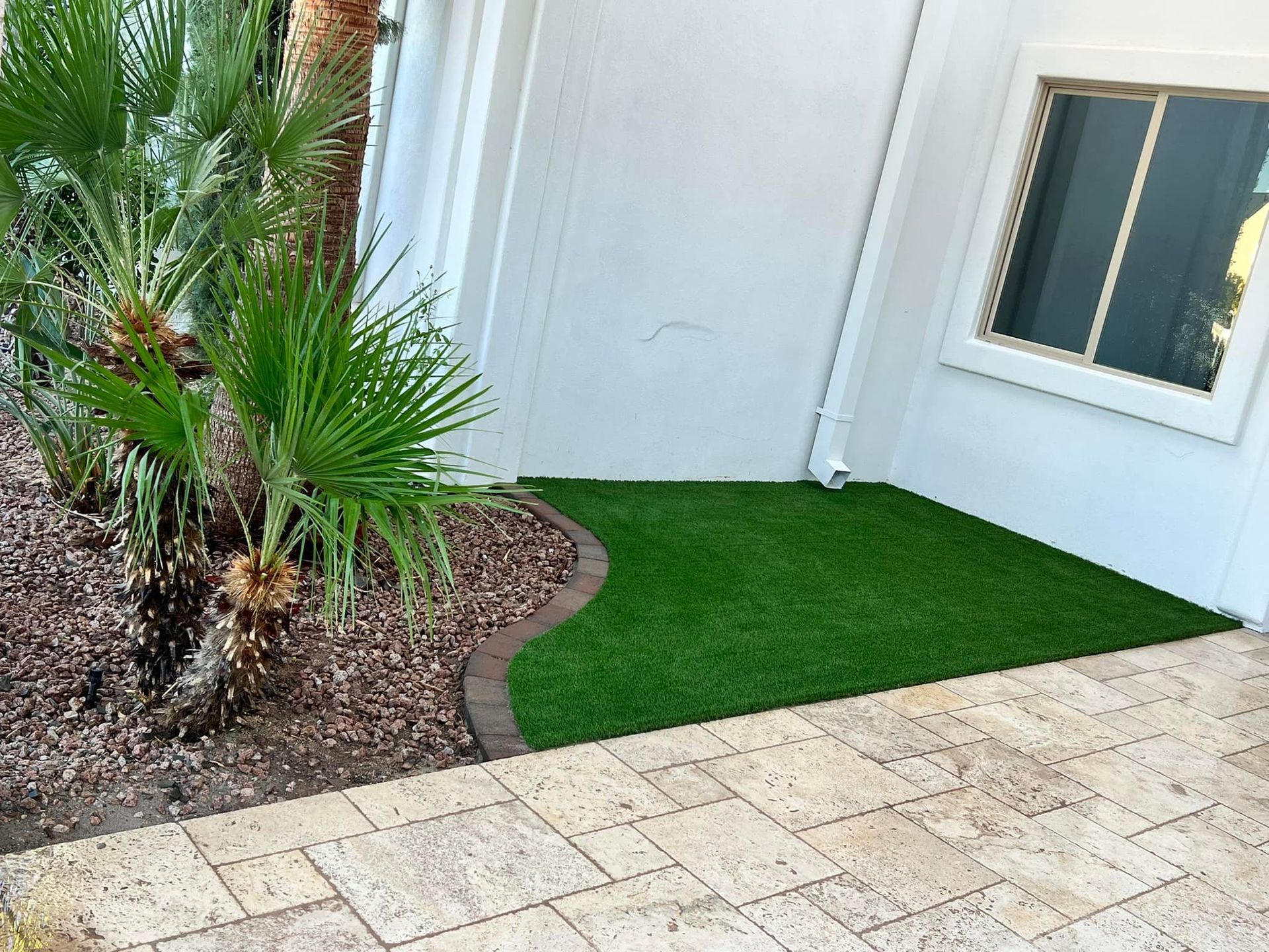 A corner of a white house with a patch of artificial green grass, surrounded by decorative rocks and stone pavers.