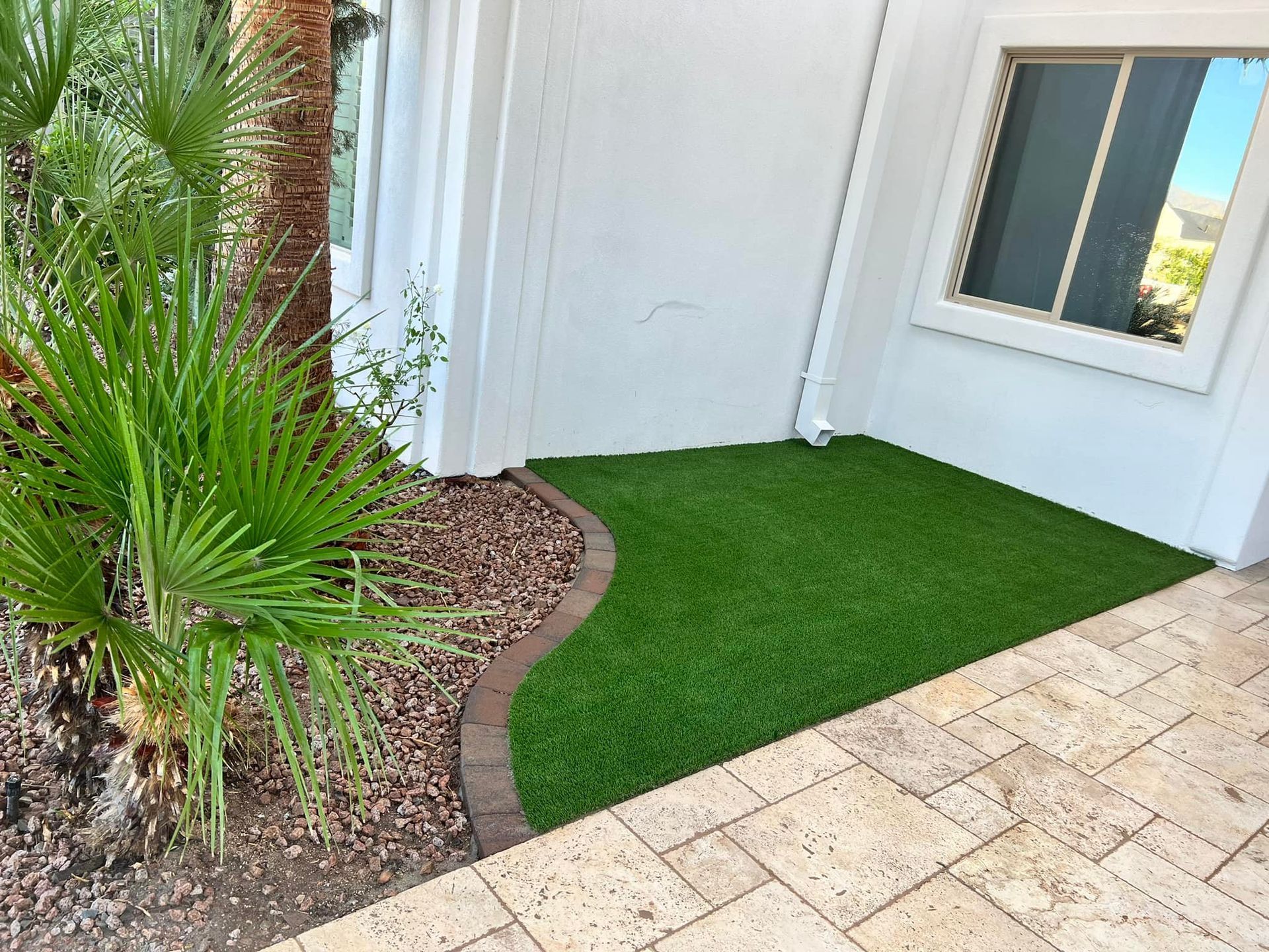 A small patch of artificial turf bordered by pavers and gravel next to a white house wall and a palm tree.