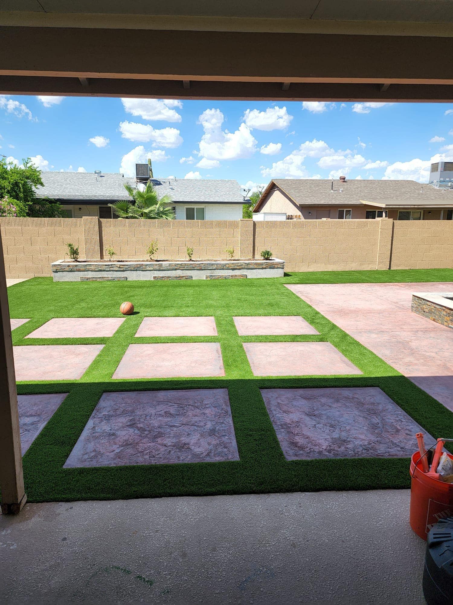 A backyard view featuring a lawn with square stone pavers, a low retaining wall, and houses under a partly cloudy sky.