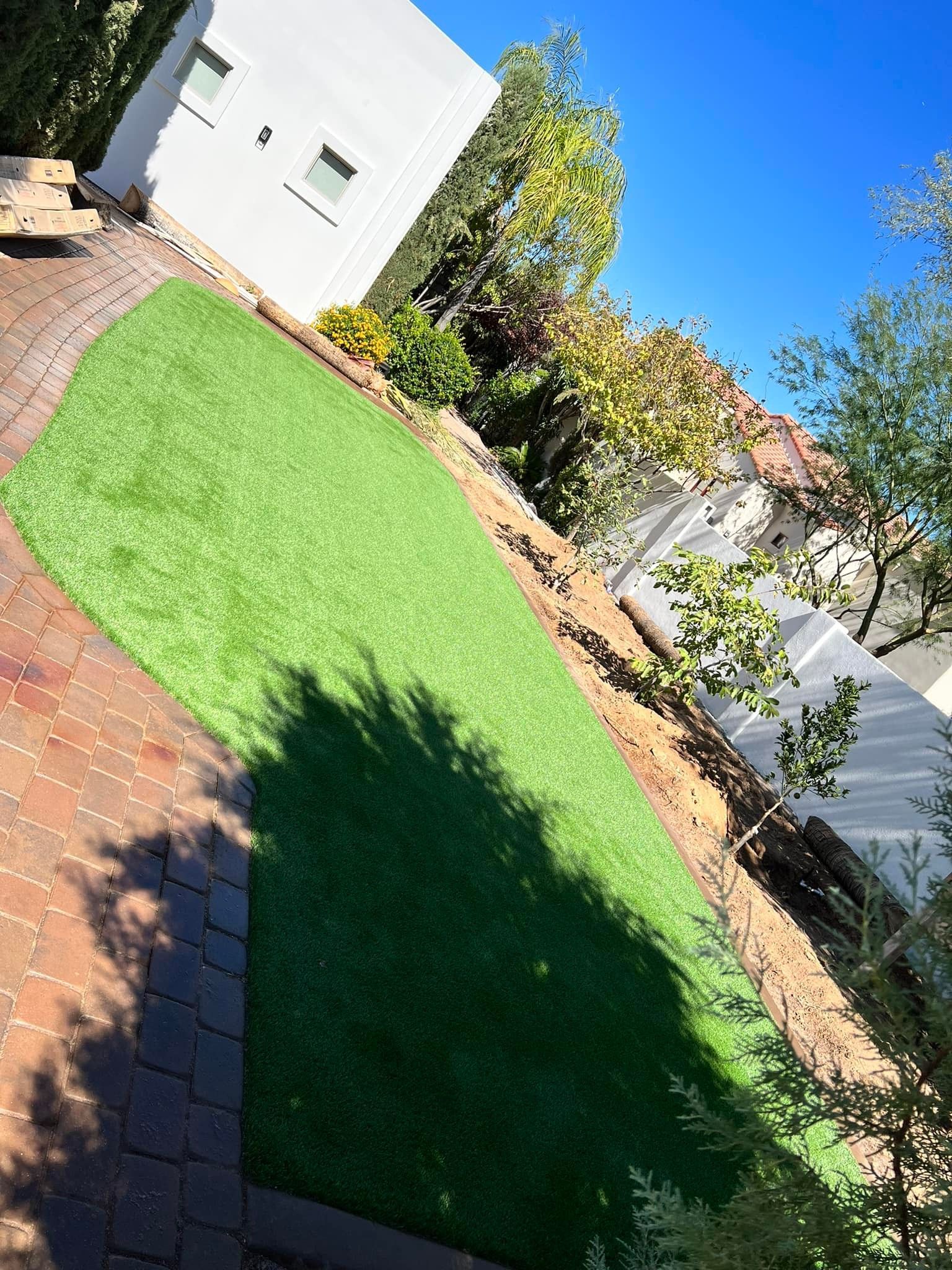 A bright green artificial lawn sits next to a brick patio, bordered by trees and a white wall under a clear blue sky.