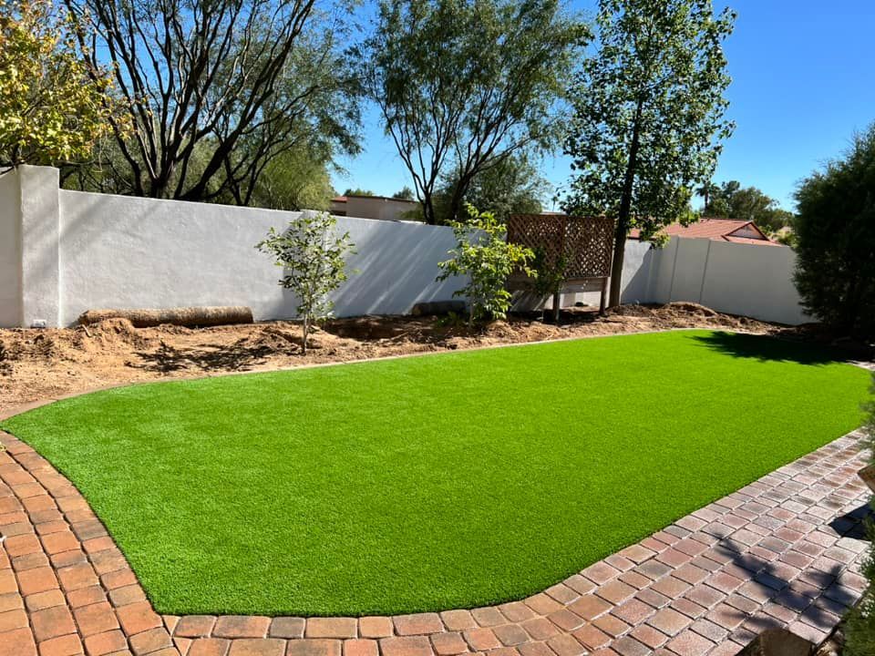 A rectangular patch of vibrant green artificial grass framed by decorative red brick pavers in a bright, sunny backyard.