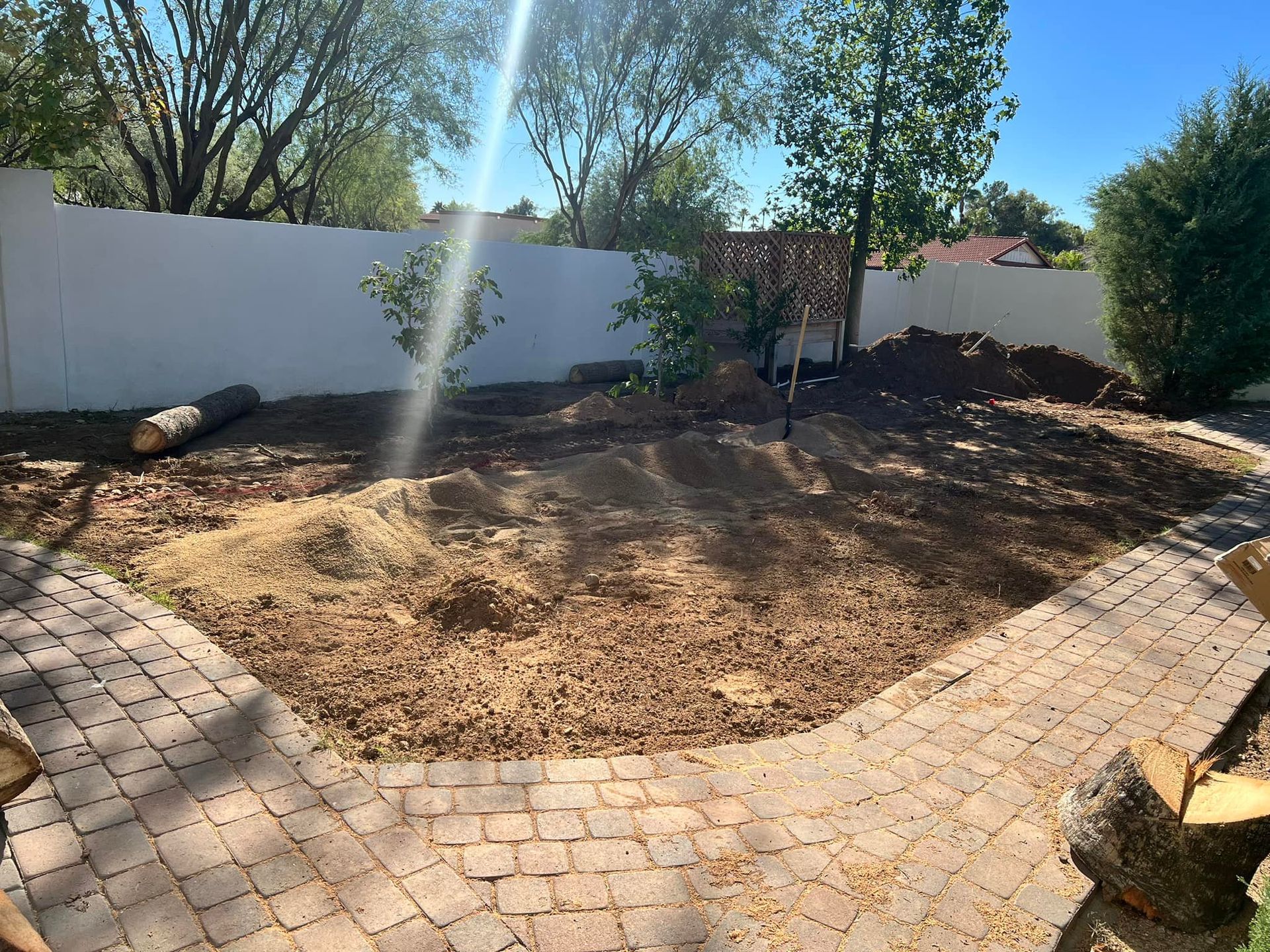 A sunny backyard with a freshly cleared dirt patch framed by brick pavers, a white wall, and sparse greenery.