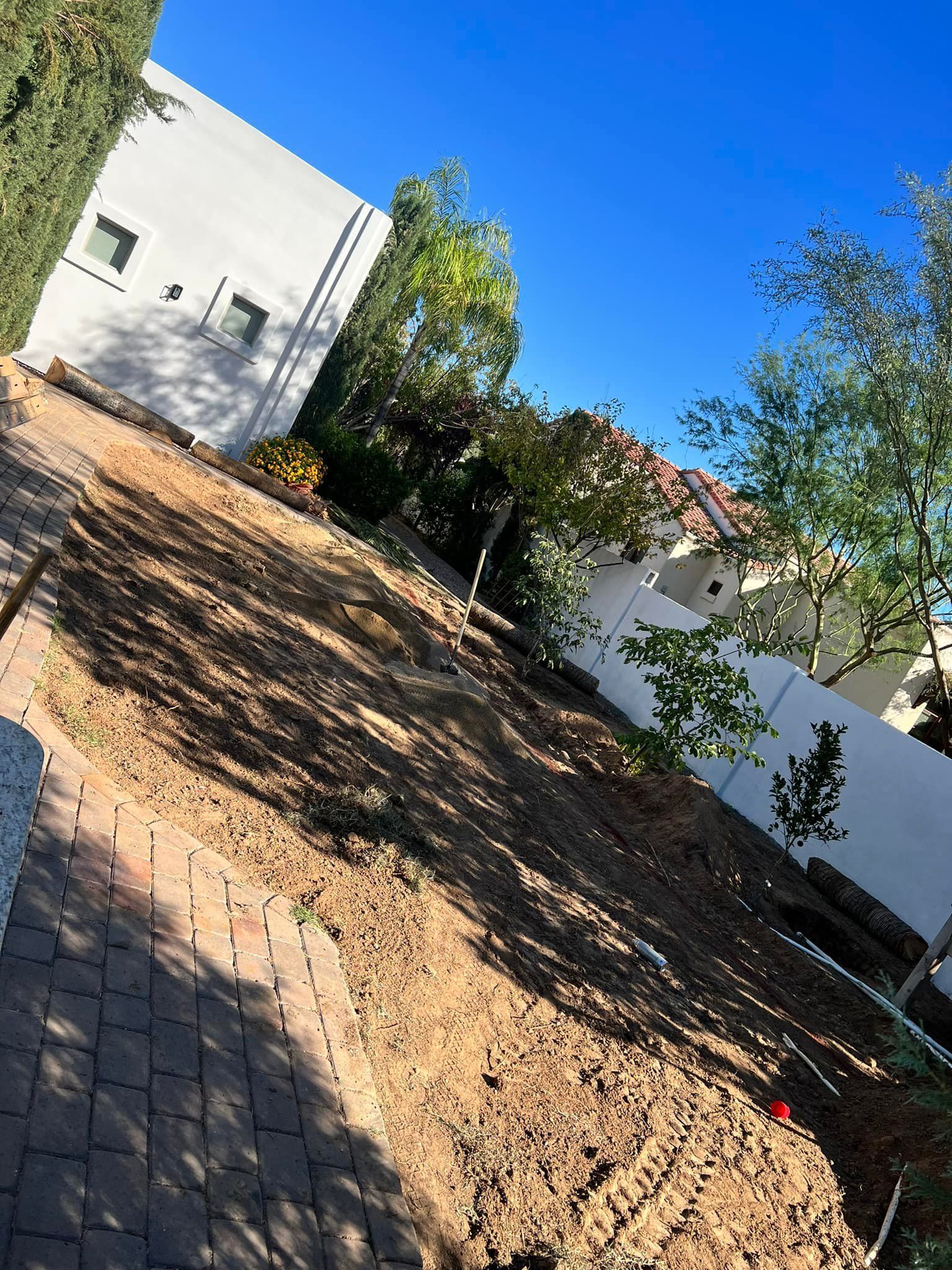 A low-angle view of a sloped, bare dirt backyard next to a white house with a brick path in the foreground.