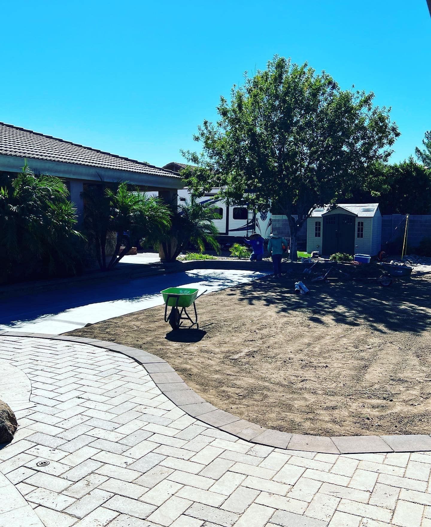 A backyard under a blue sky featuring a paved stone walkway, a patch of dirt, a green wheelbarrow, and a small shed.