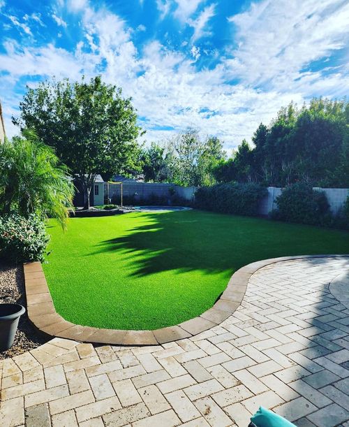 A manicured green lawn bordered by a curved stone wall and paver patio under a bright blue sky with scattered clouds.