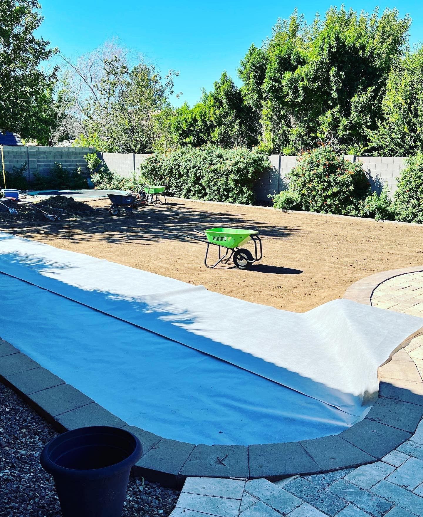 A backyard renovation showing a partially covered swimming pool, a dirt yard, and a green wheelbarrow under sunny skies.