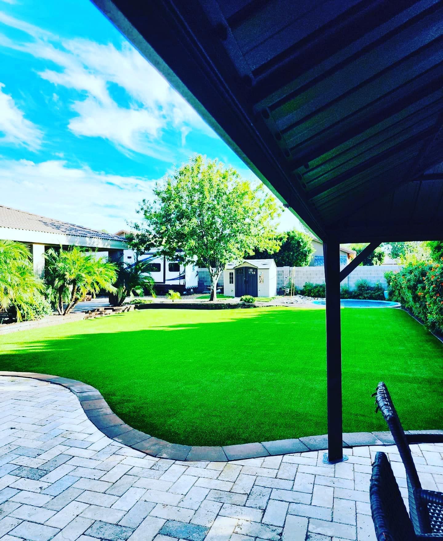 A backyard view featuring a bright green lawn, a brick patio, a covered patio area, and a large tree under a blue sky.