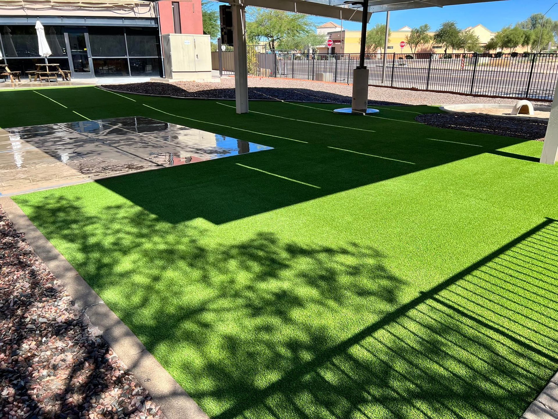 A sunny outdoor patio with bright green artificial turf, a reflective table, and support pillars under a shaded structure.