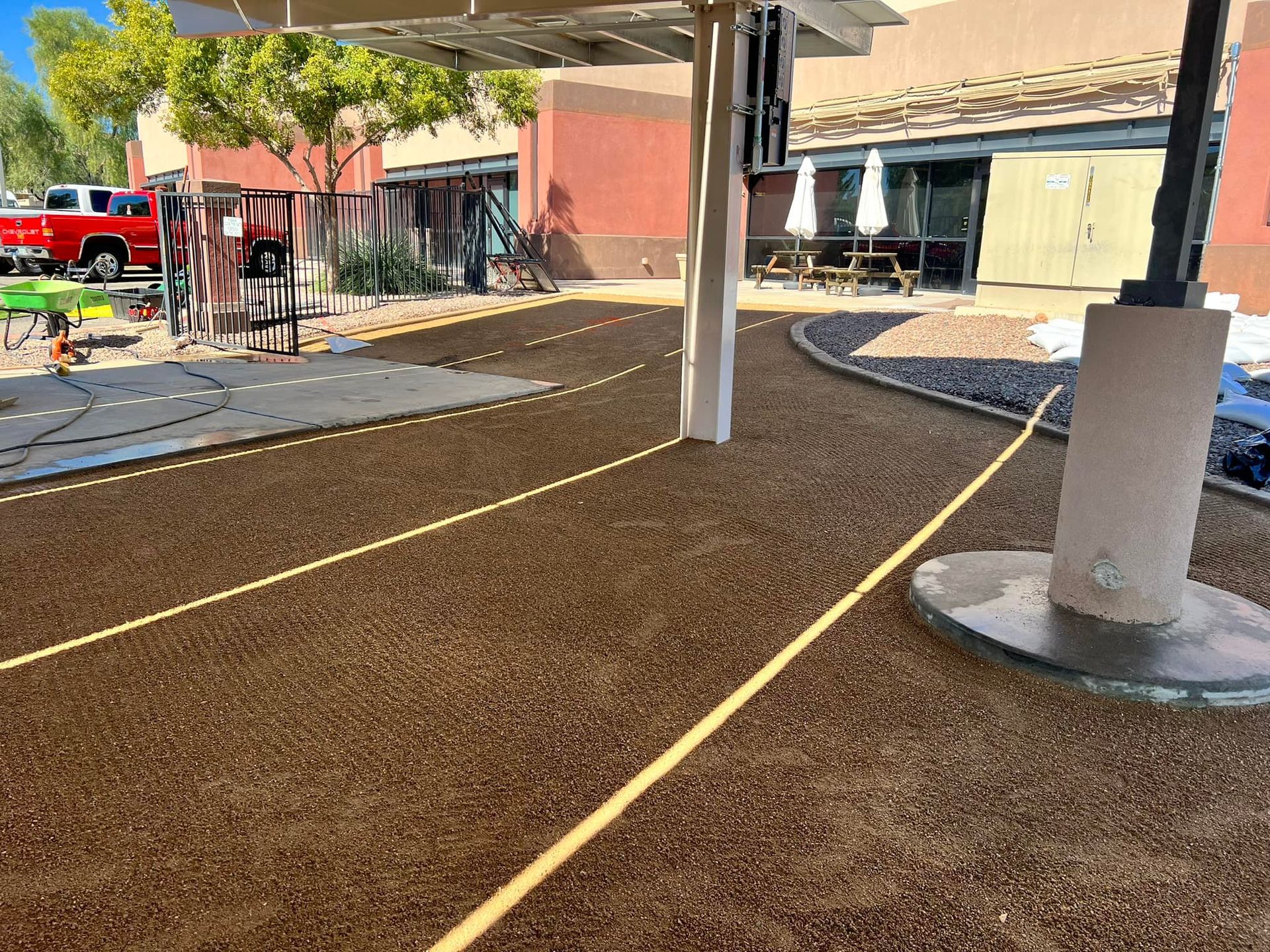 A gravel area under a solar shade structure with yellow string lines marking a path, next to a building and red truck.