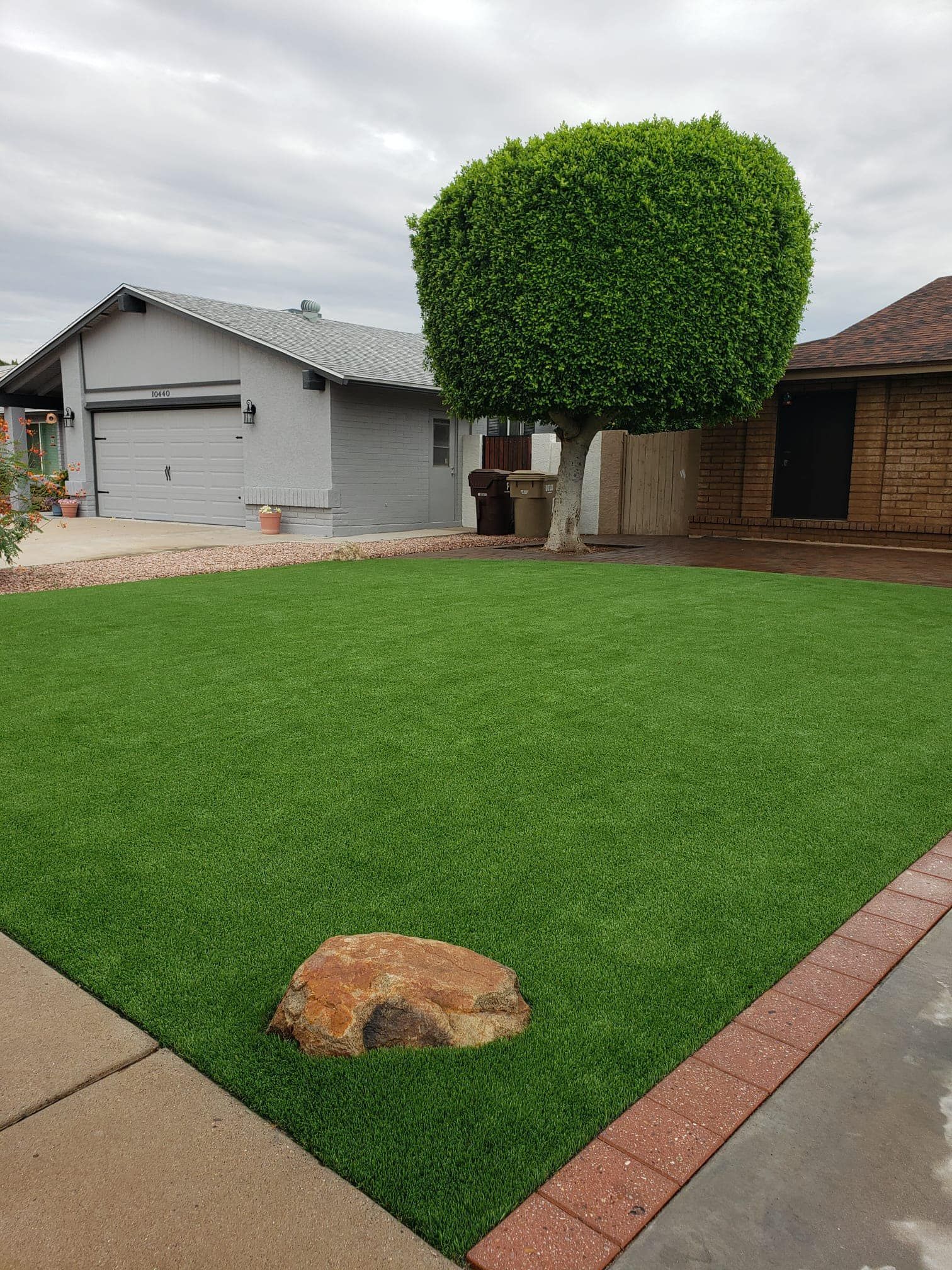 A manicured green lawn with a large rock in the foreground, a round-top tree, and a garage in the background.
