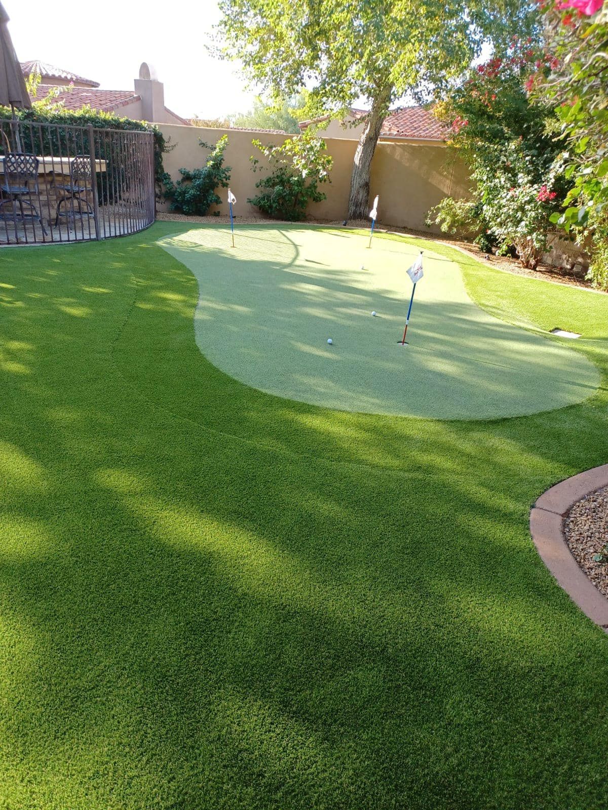 A backyard putting green with two holes and flags set in a neatly manicured lawn, bordered by landscaping and a fence.